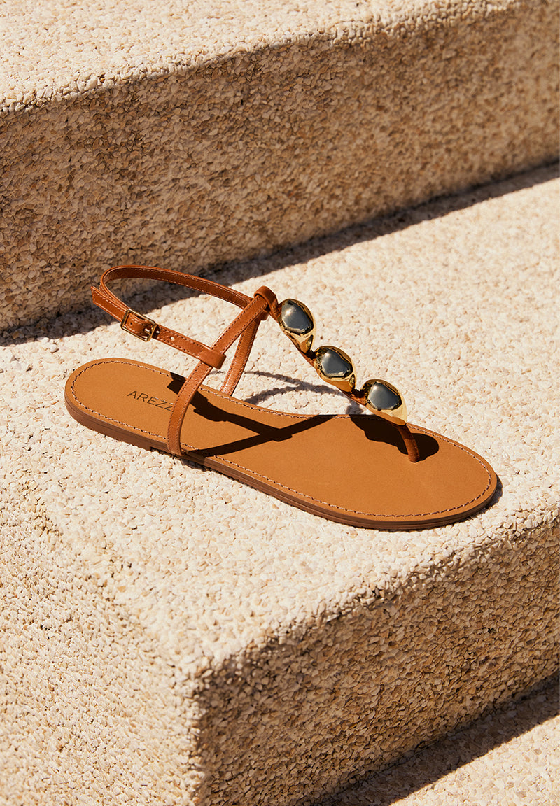 Promotional the Soroya Flat Sandal, a Brown leather sandal. The style features a flat sole and a rounded toe. It has a thin strap that runs between the toes, adorned with bold, organic-shaped gold accents, connected to another thin horizontal strap. Plus, it includes a thin ankle strap with a metallic side buckle closure. The insole is brown with the brand name engraved. 